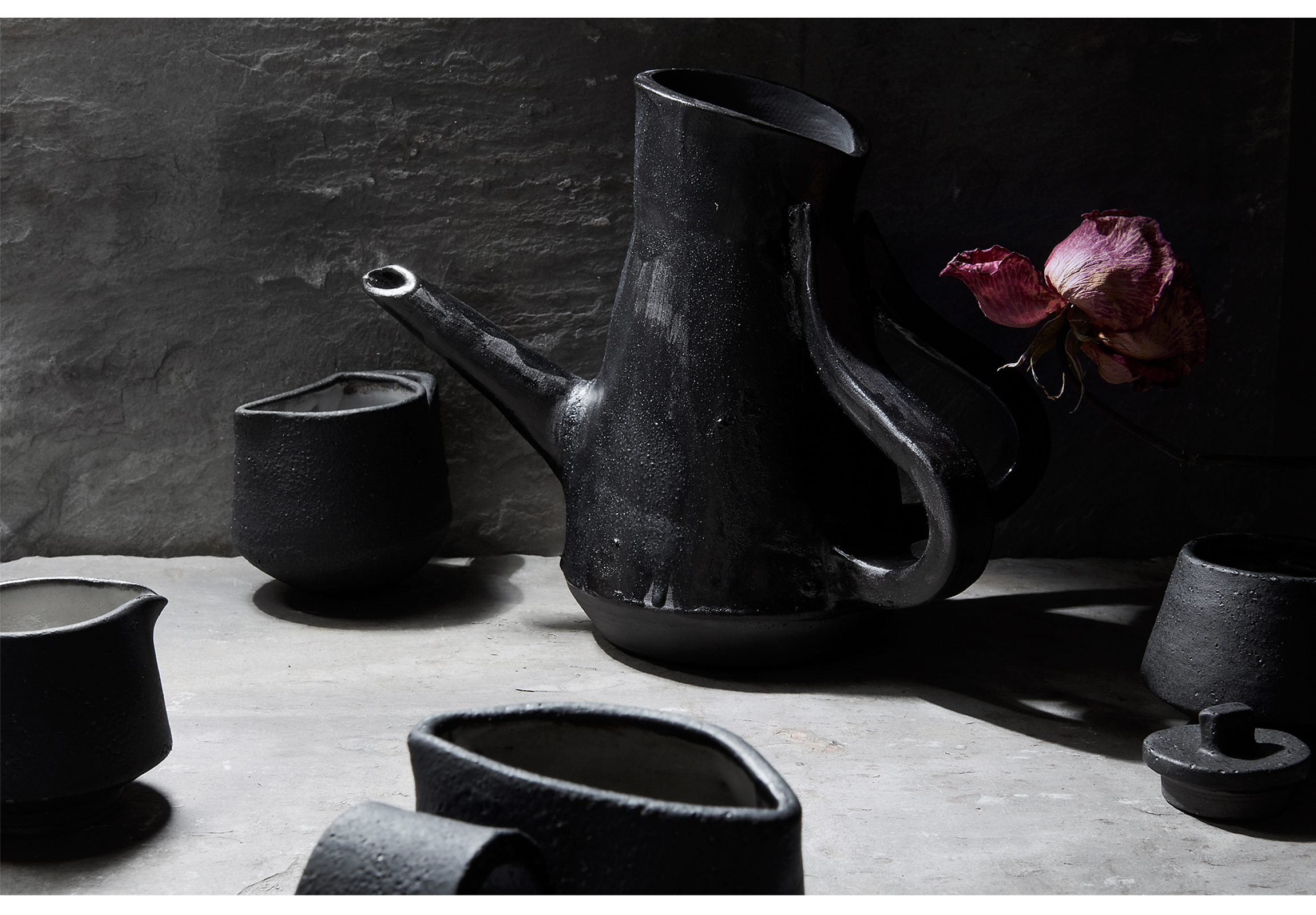 Black glazed tea set on white table with black background and a purple flower