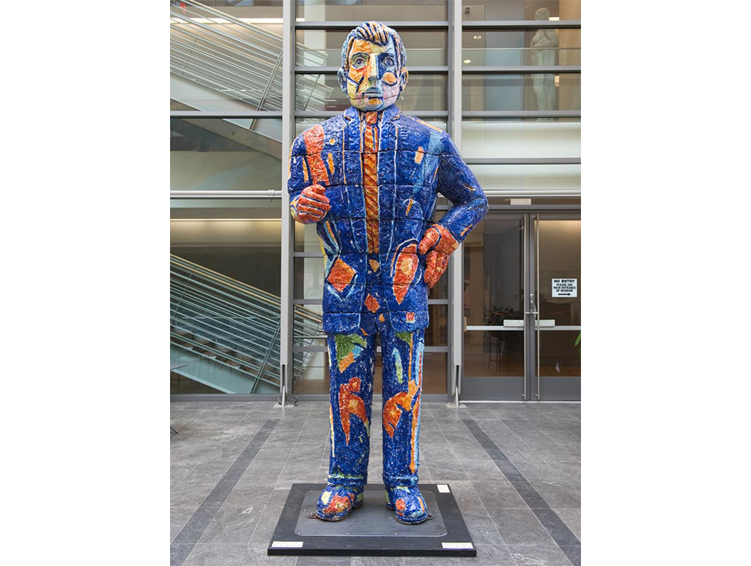 Colorful blue and red brightly colored 10 foot sculpture of a man, placed on a peastal on a grey stone floor with glass windows in the background