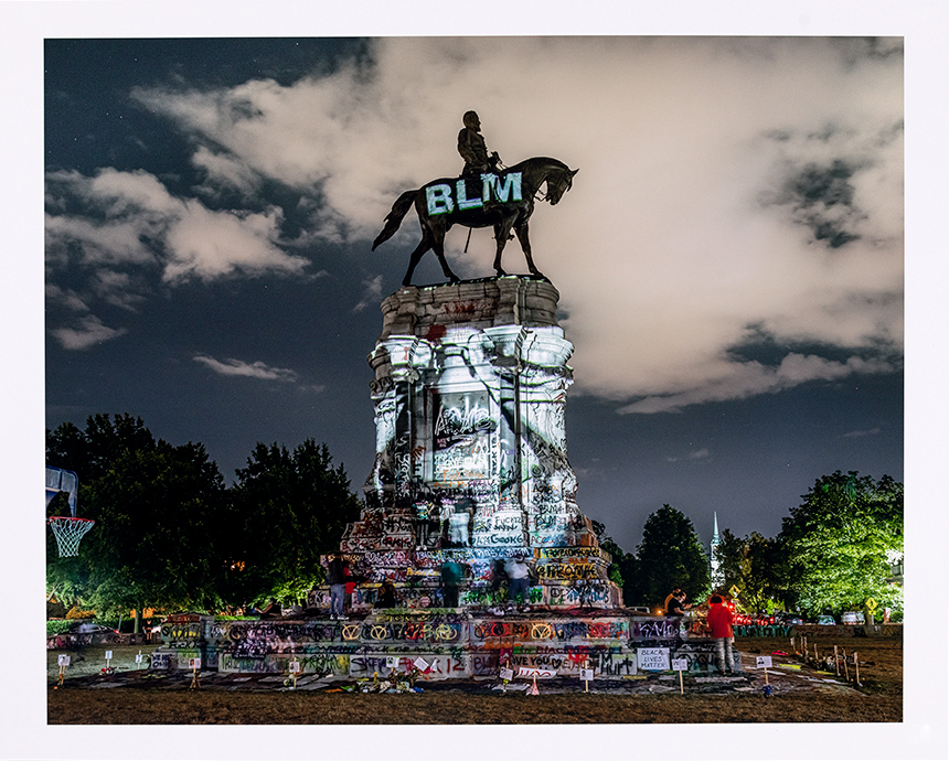 American monument with colorful projection reflected on it with BLM on the silhouetted sculpture of a person on a horse at the top, all at dusk