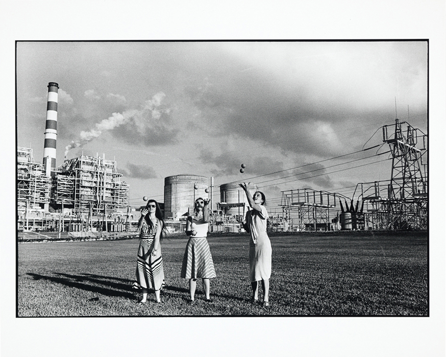 Black and white photo of three people juggling in dresses in front of a nuclear power plant