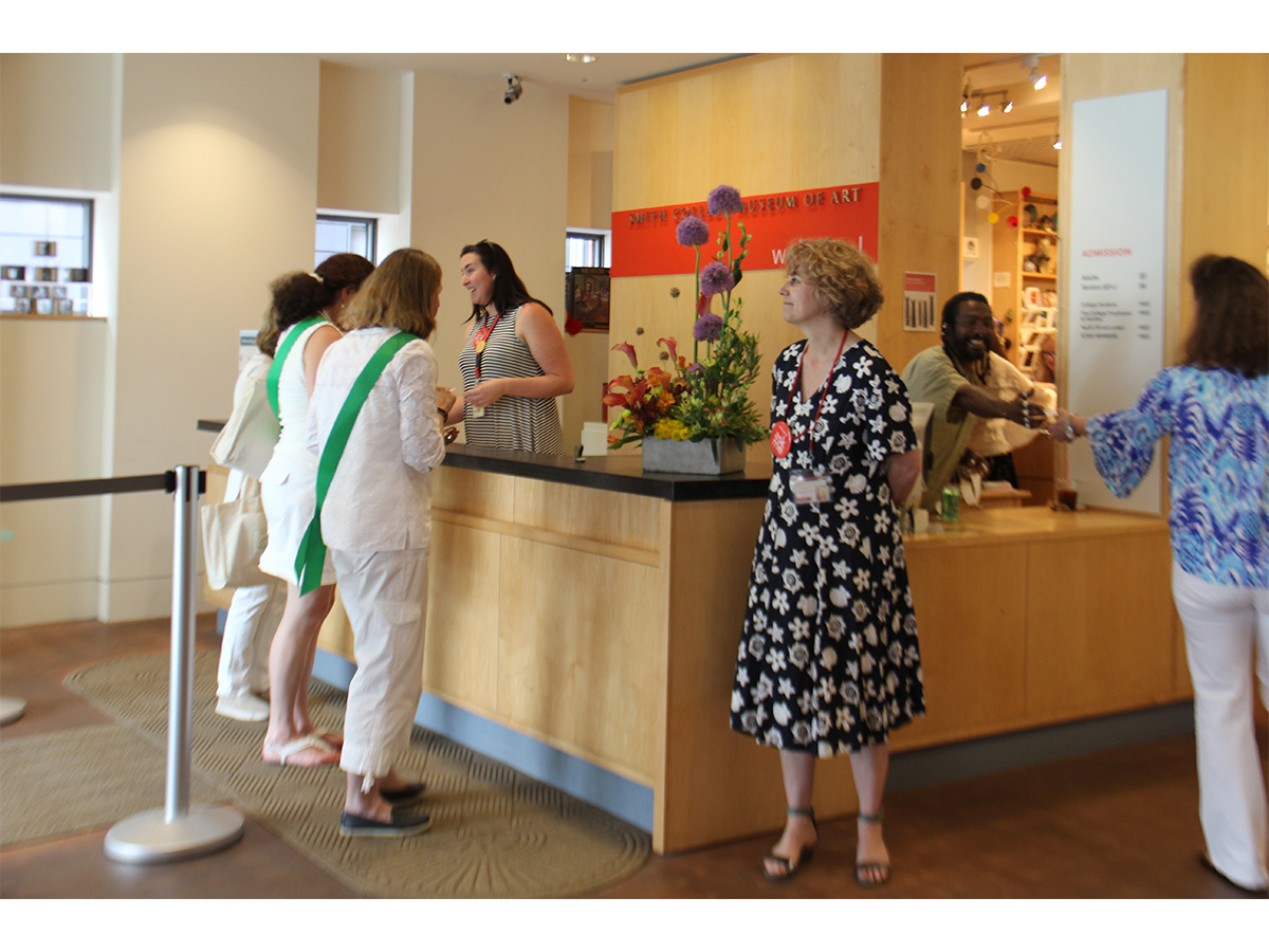 Three Smith alumnae visitors at the front of the admissions desk with two staff behind desk, one interacting with a visitor to the right of the desk and another staff member standing next to the desk ready to greet visitors