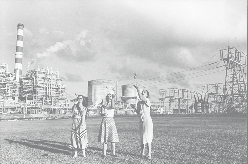 Black and white photo of three women juggling in front a nuclear power plant