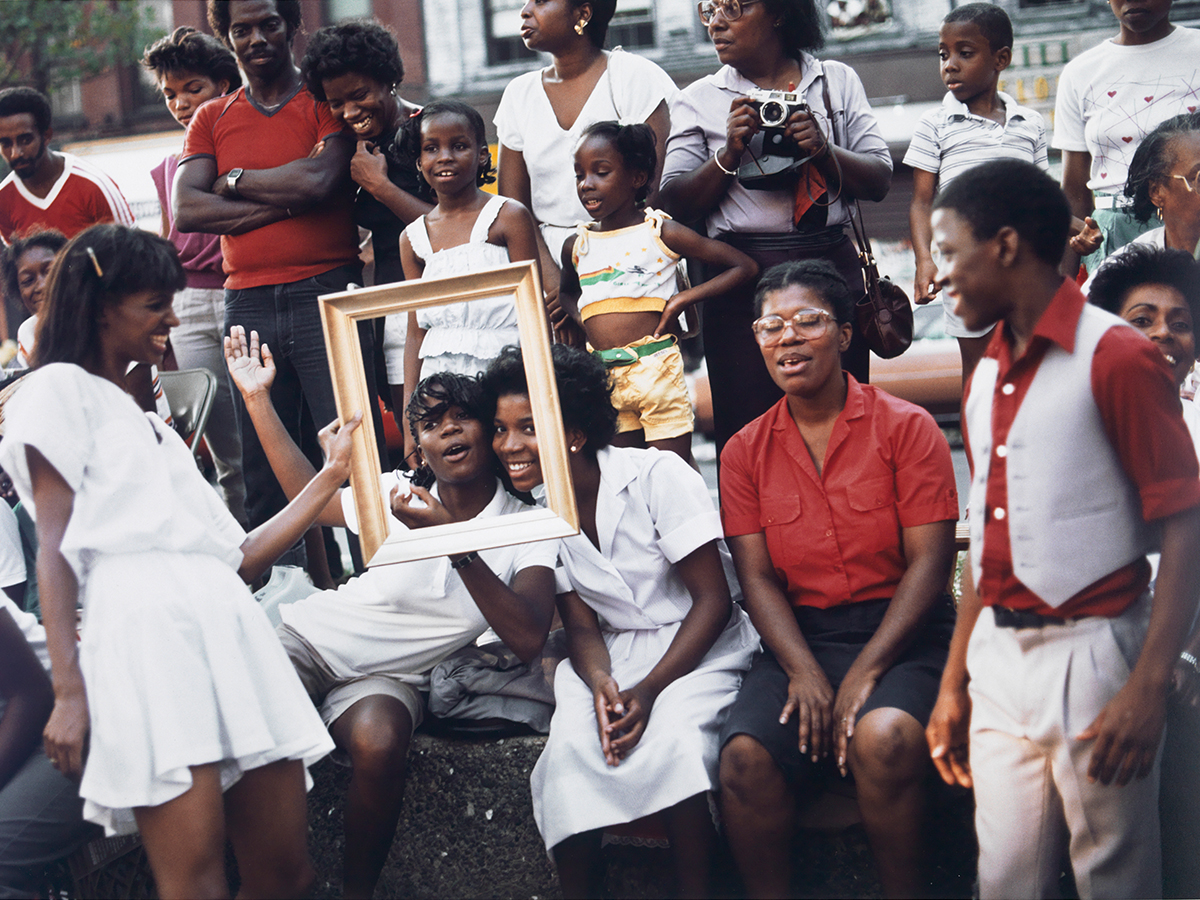 Photo of a crowd of people of all ages from 70's, dressed in white and red, with one woman holding up a frame in front of two people's faces