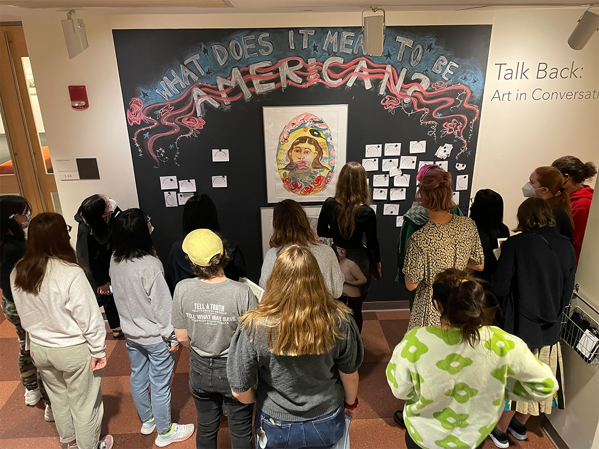 Photo of students looking an artwork hung on a chalk wall with notes posted beside the art and chalk on the wall above the artwork