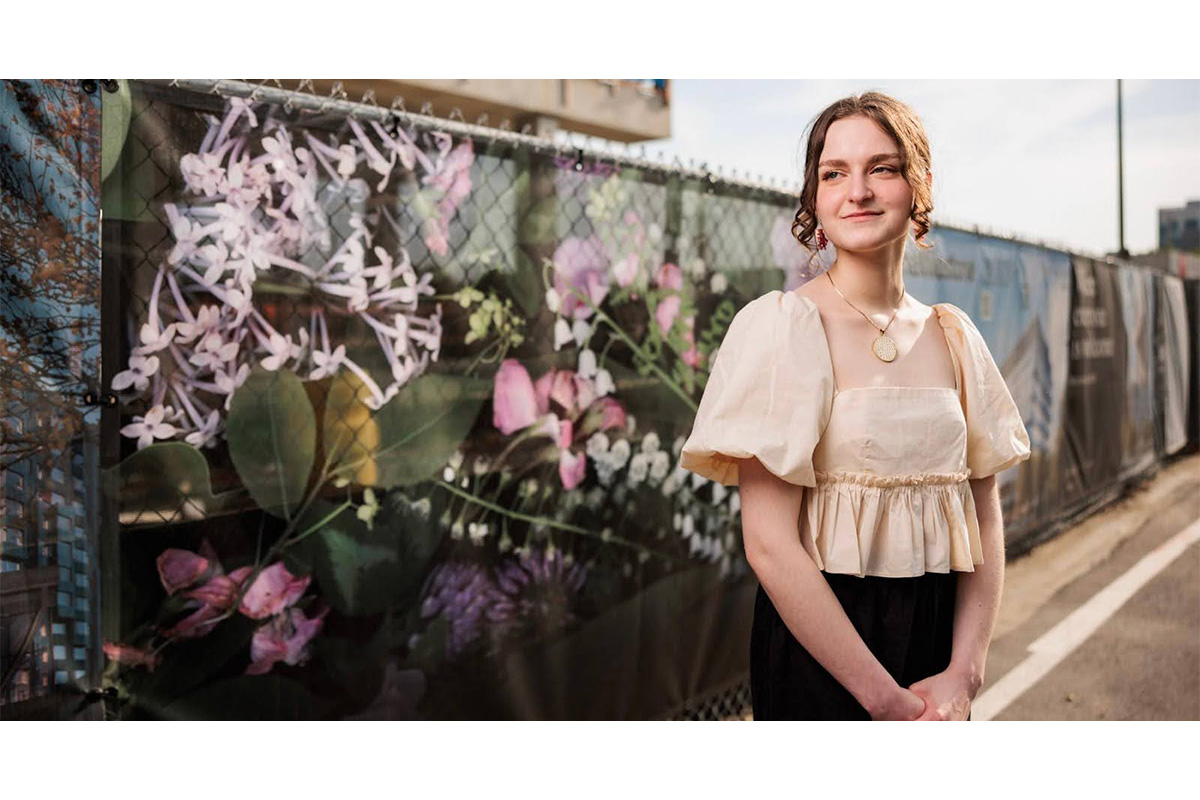 Three-quarter portrait of Grace standing in front of her artwork – photos of flowers on a dark background – displayed on a fence as part of the Harvard Public Art & Culture Tour.