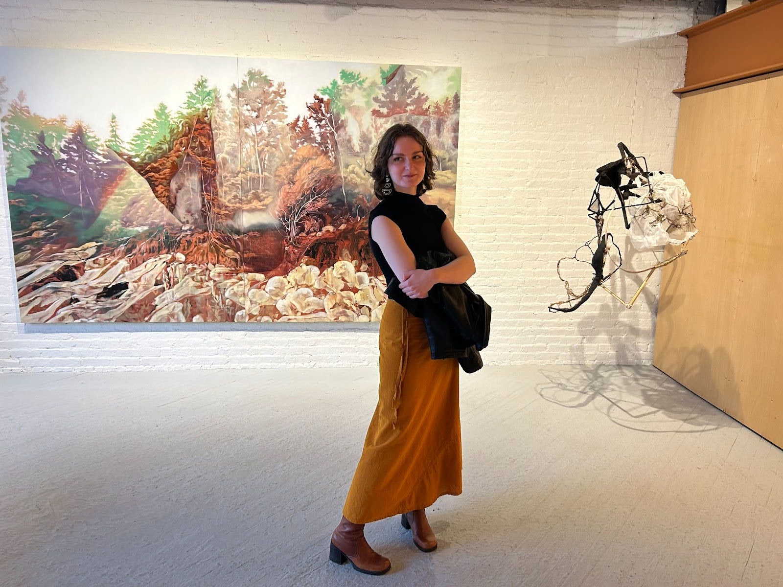 Grace standing in a long yellow skirt and black top in front of a landscape painting and black and white hanging sculpture in a gallery space.