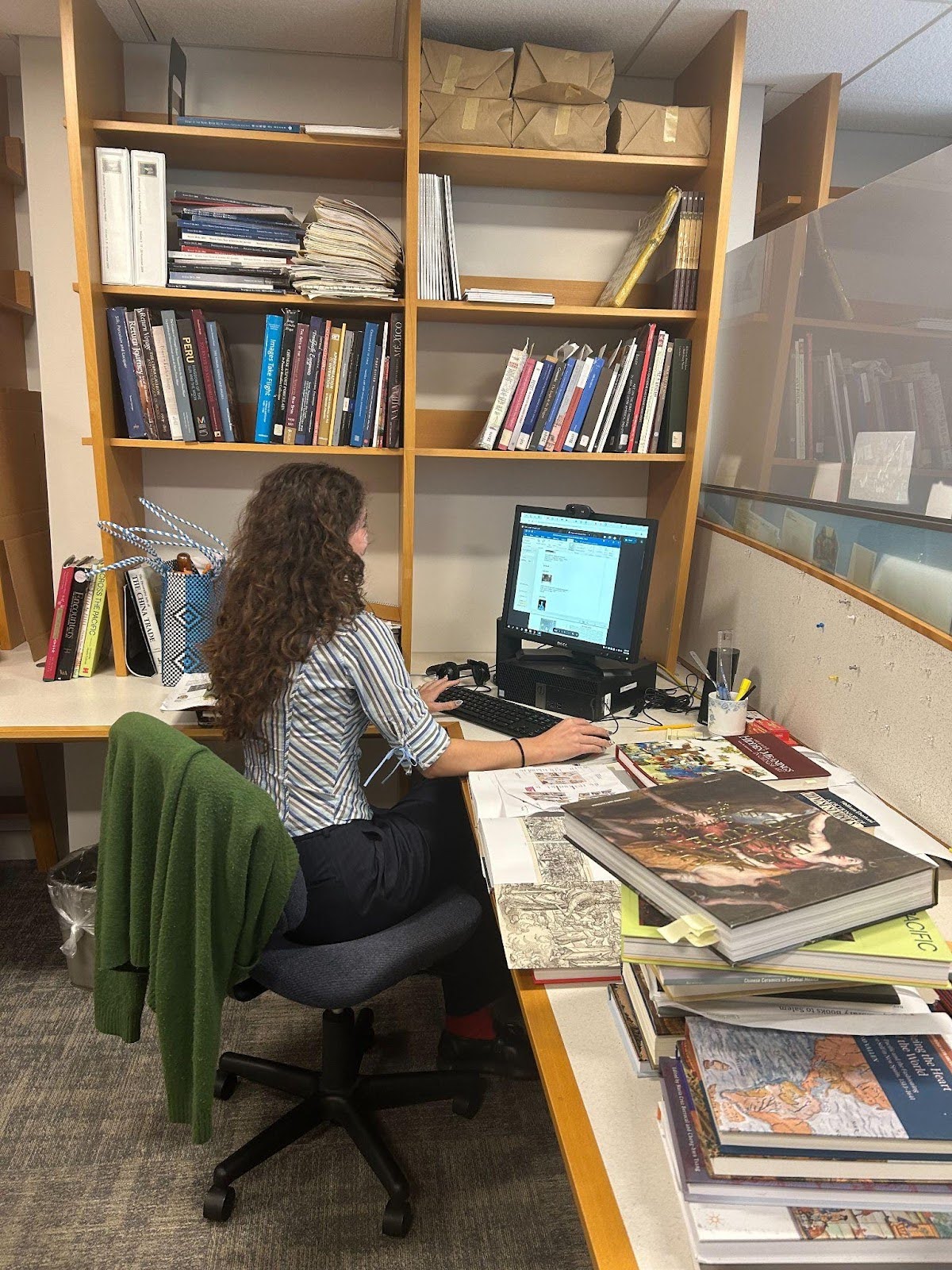 View of Lucy from behind sitting at a desk, typing on a computer and surrounded by books.