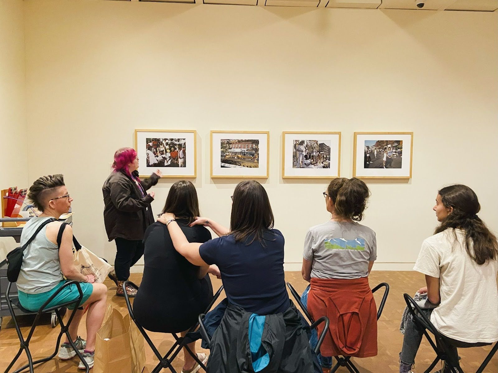 Lilly gesturing towards four photographs on a gallery wall as a group of five visitors sit in the foreground and look towards them.