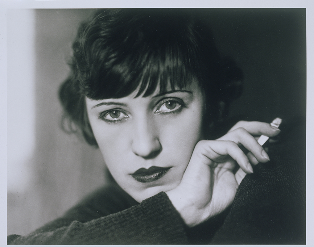 Crisp black-and-white photograph portrait of a woman with one of her hands holding a cigarette diagonally across the composition. She looks straight into the camera and has dark, bobbed hair and dark eyeliner and lipstick.