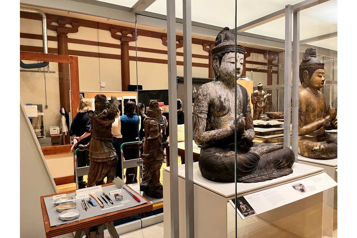 "gallery containing several statues of buddha with students wandering around"