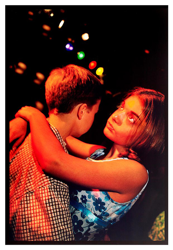 "dark interior with colored lights, close-up of very young couple dancing, boy wearing a checked shirt and girl and sleeveless blue flowered dress"