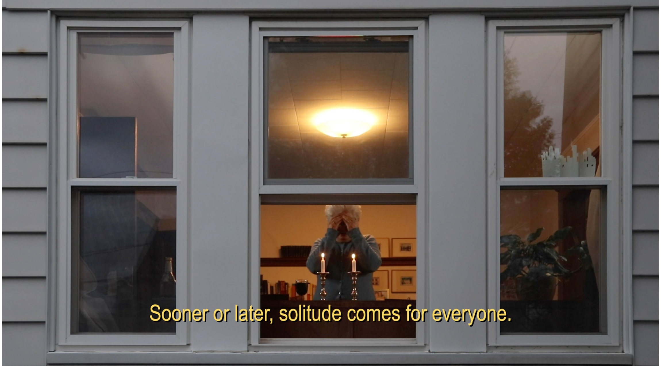 woman seen through windows of house, standing in front of candles. text at bottom reads: "Sooner or later, solitude comes for everyone."