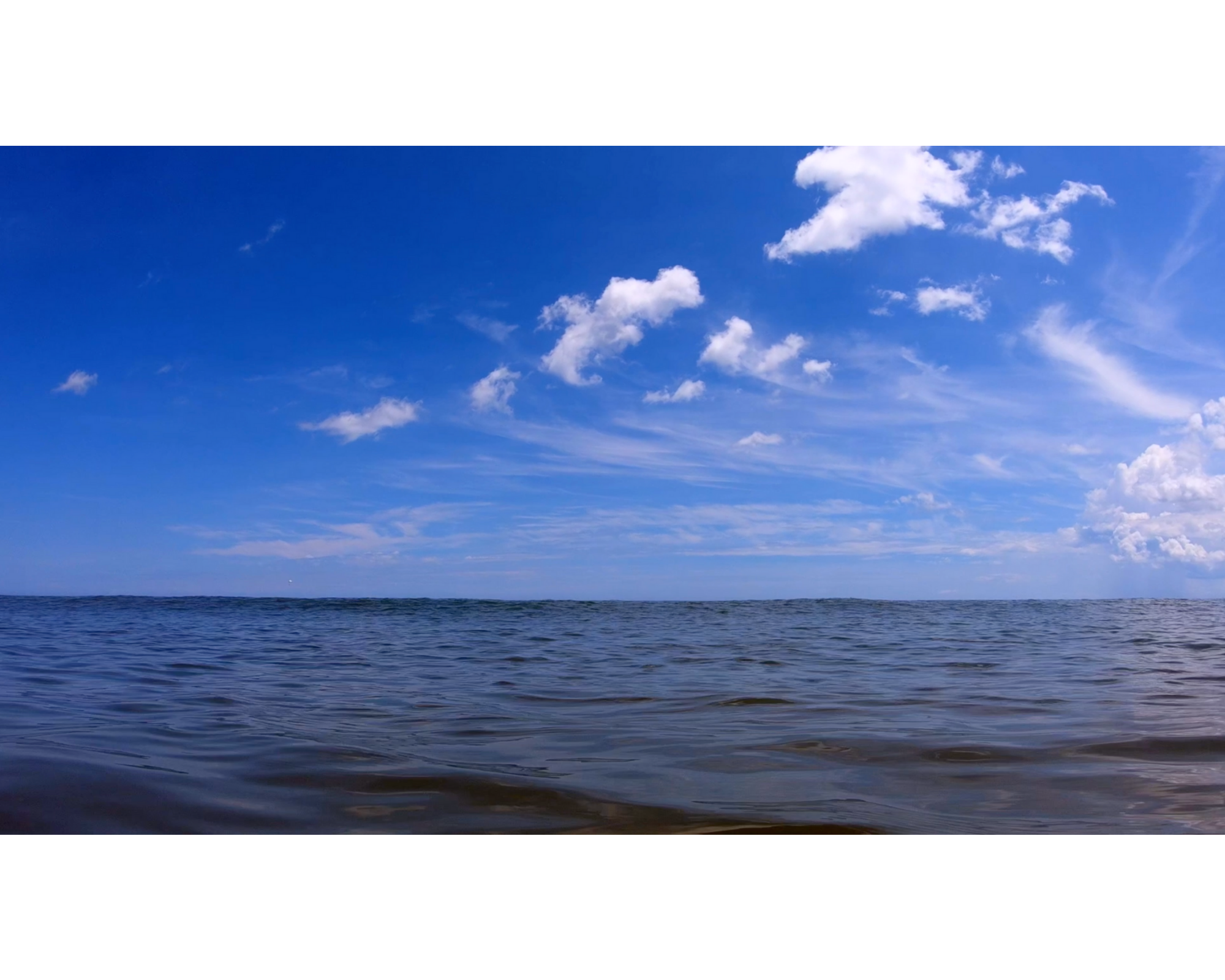 Still from Hopinka's "Cloudless Blue Egress of Summer": slightly cloudy blue sky filling top of frame with ocean water below the horizon line.