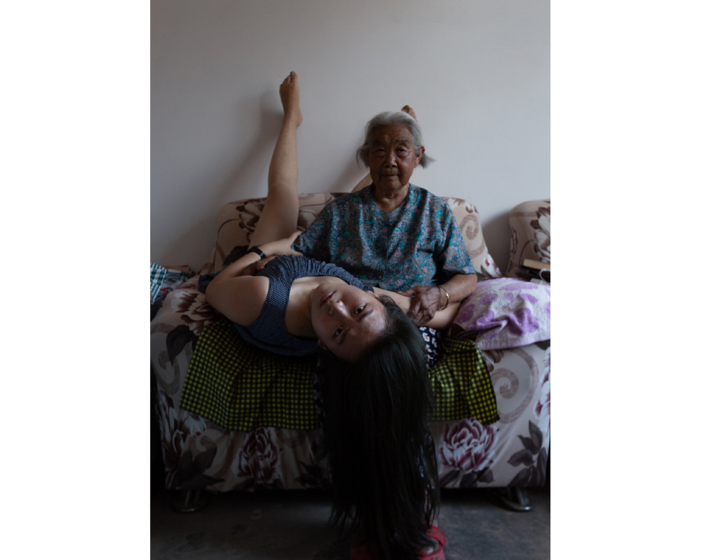 Photograph shows a young women looking upside down at camera. Her legs and feet are up against a wall. She is lying next to an older woman who is sitting straight up in the large upholstered chair covered in patterned textiles. They are holding each others arms.