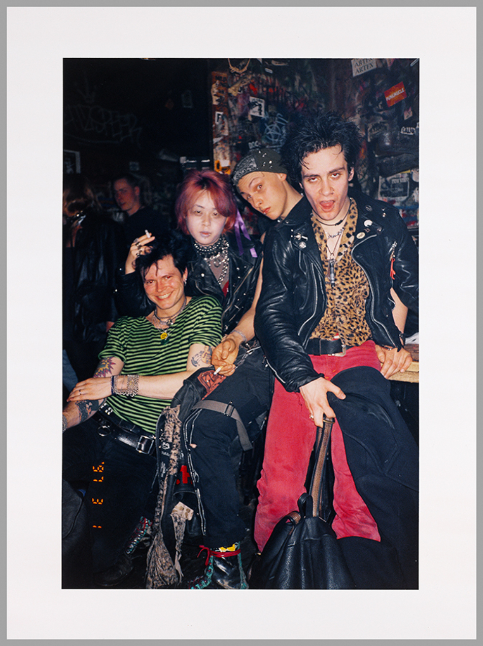 Four seated people with tattoos and piercings dressed in leather, stripes, and animal prints looking directly at the camera.