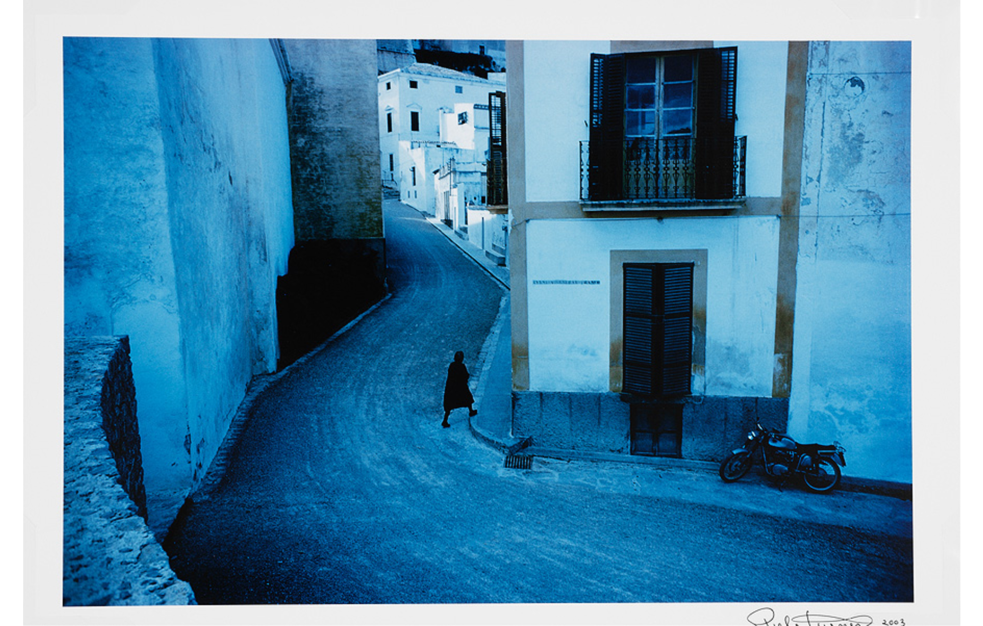 Overall blue tone; curving village street, plain building walls up left side, motorcycle parked near closed windows at lower left, woman in black stepping onto sidewalk at lower center which follows road into picture past small whitewashed box-like houses.