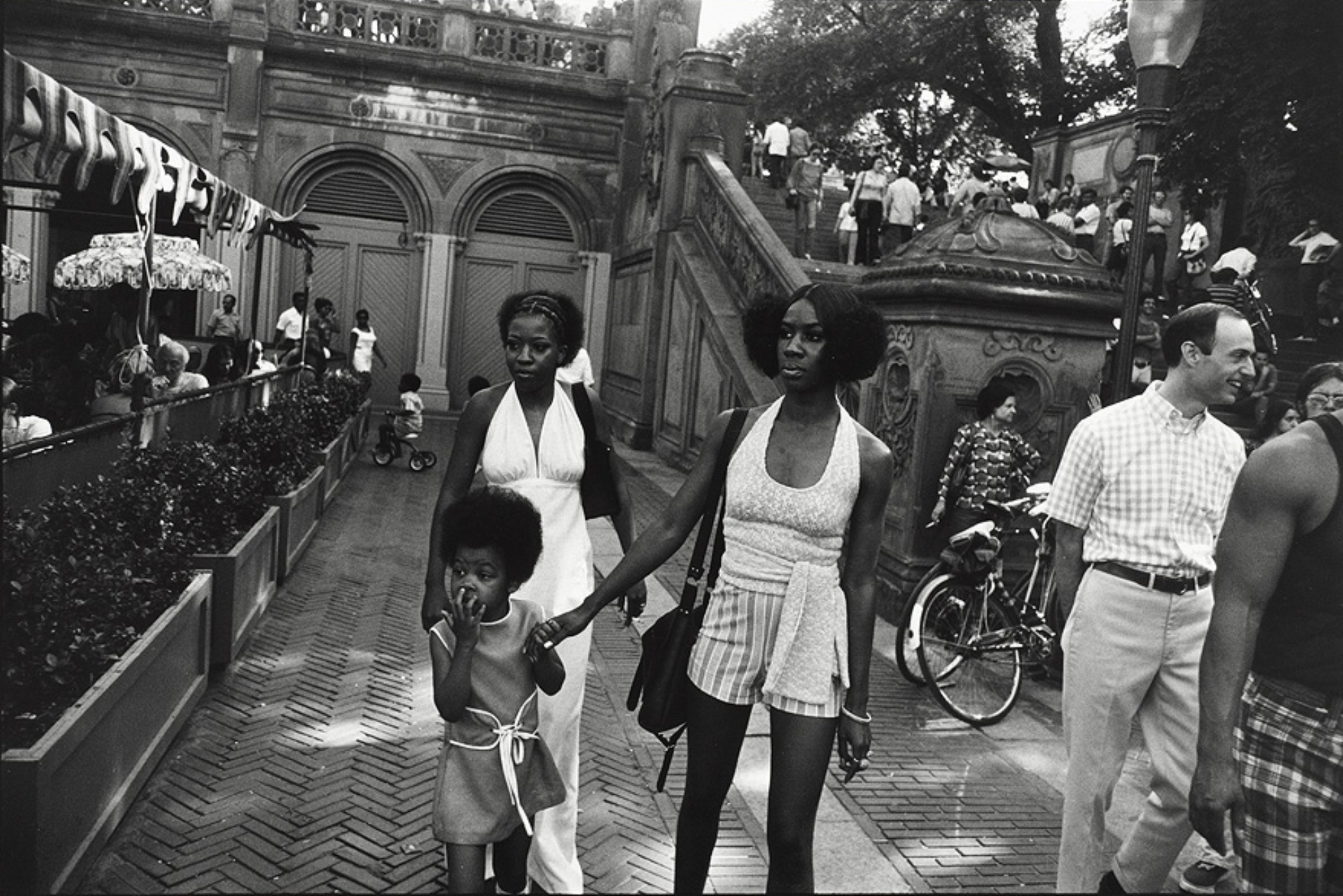 Two black women, one holding child's hand, walking away from stone stairway, restaurant on left and people on right.
