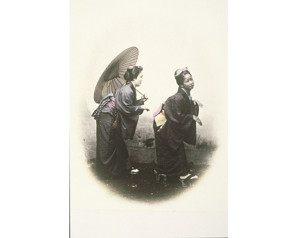 two Asian women, dressed in kimonos; one carries a parasol 