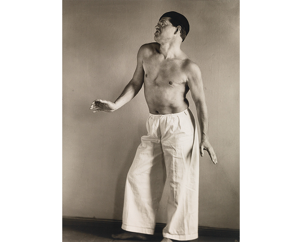 man standing with bent knee, raised shoulder, and flexed hand, wearing white pants and no shirt