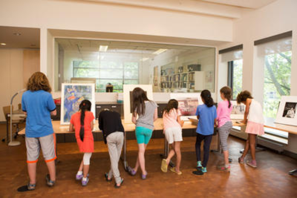 Young students in the Cunningham Center gallery looking at works of art on tables; from behind.