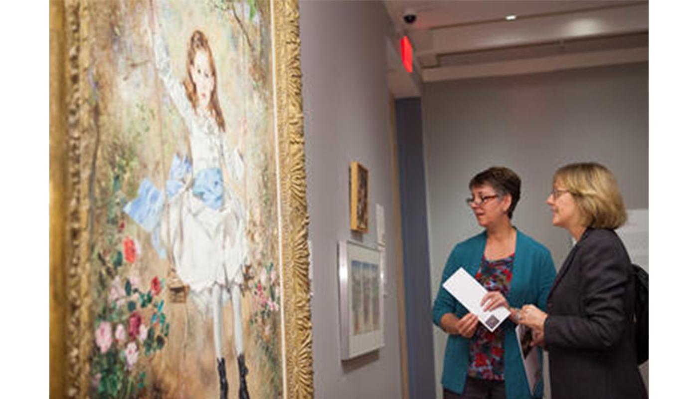 two women stand in front of a painting of a girl on a swing, looking at the painting and talking to one another