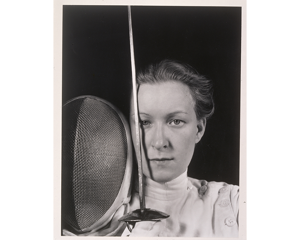 bust portrait of woman in fencing uniform, holding sword vertical over right eye and mask beside that