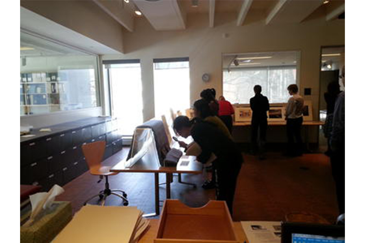 tables set up around a room with artworks propped up on them. students look closely at the artworks using magnifying glasses