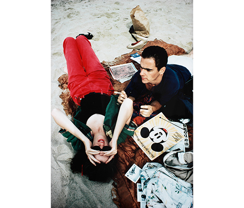 close-up of woman lying on her back on a rust colored quilt on a beach, she has dark hair, holds her sunglasses on her face with both hands, and wears red jeans, black leotard and green shirt, next to her is a man with short dark hair, lying on his stomach, holding a cigarette in his proper left hand, he has on a blue T-shirt and white shorts