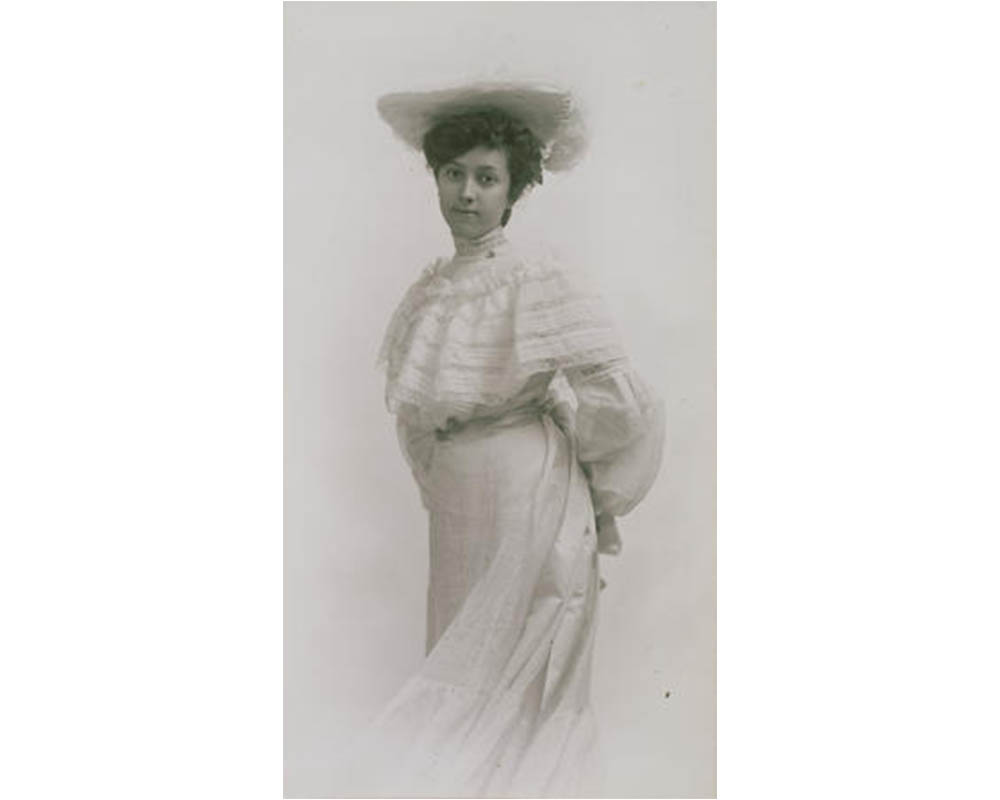 woman stands facing the viewer with her hands behind her back, wearing a fancy updo in her hair, a white hat, and a white dress