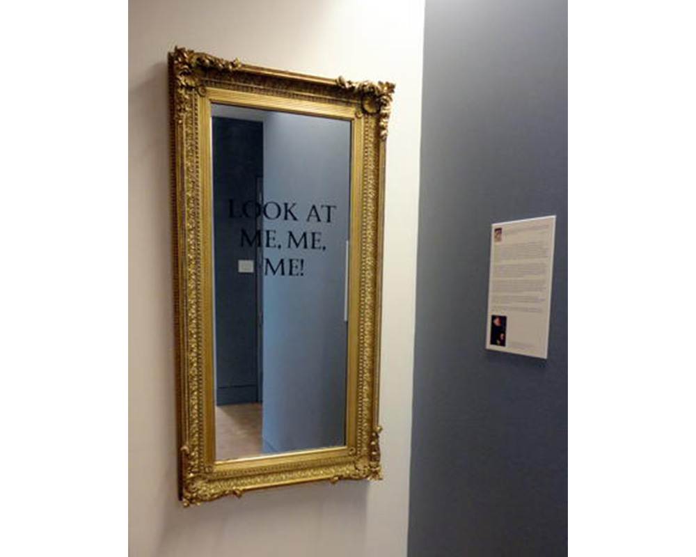 art gallery with mirror hanging on one of the walls. mirror has gold frame and black text on it that says, "LOOK AT ME, ME, ME!" 