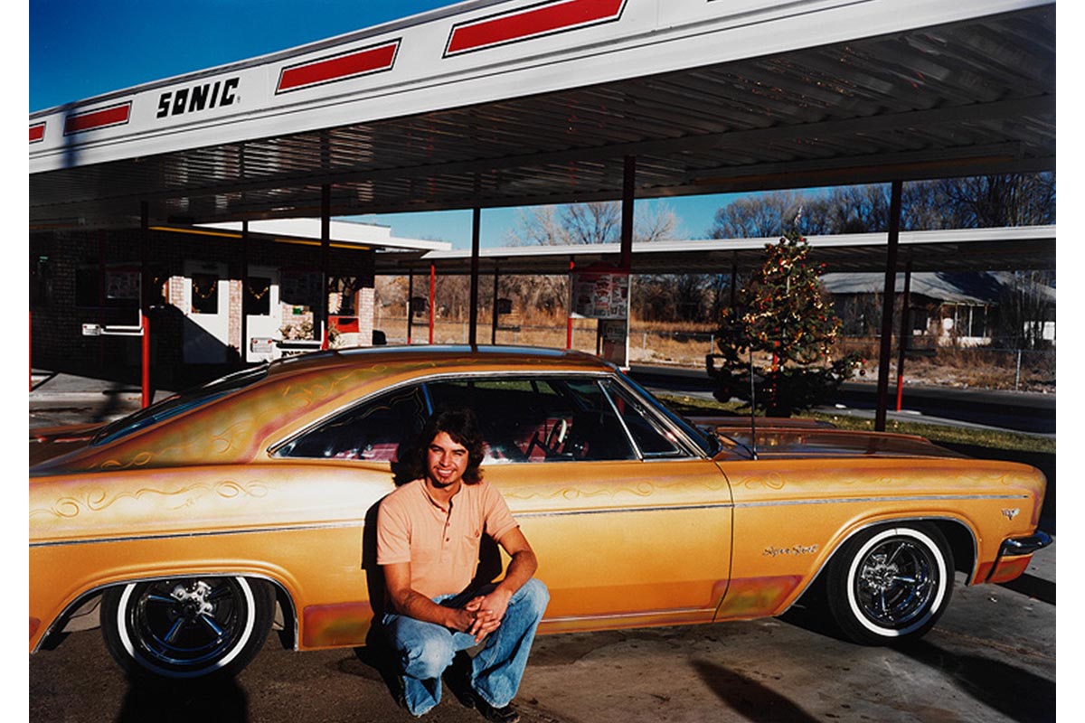 Sonic gas station with Christmas tree in background, sedan painted dull orange/gold with swirled designs, young man in jeans and short sleeve shirt squatting down near passenger door and smiling