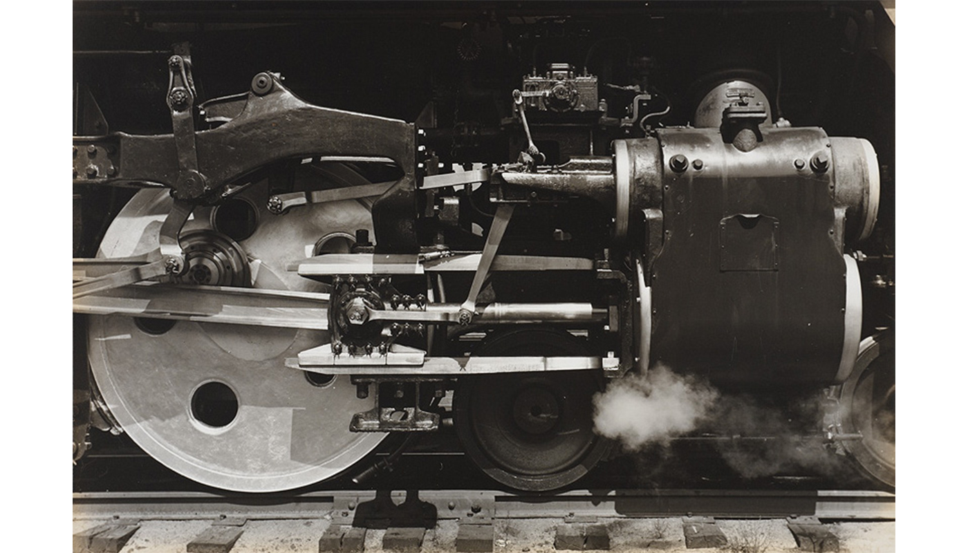 close-up of side of locomotive engine showing large wheel at left, horizontal piping, engine sections including a large box-like structure at right with steam visible underneath, section of track runs across bottom image, top image is in deep shadow