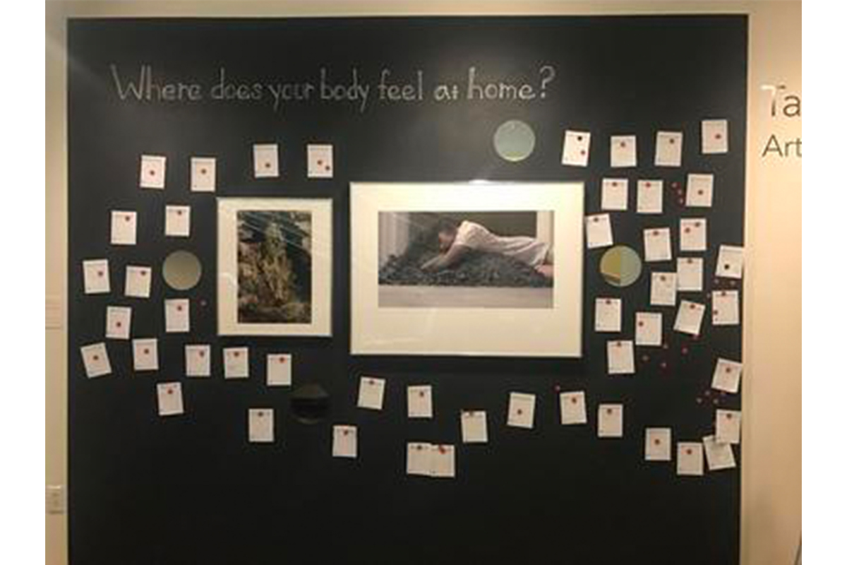 installation of two artworks against a black wall, with the question "where does your body feel at home?" written at the top and written responses tacked to the wall