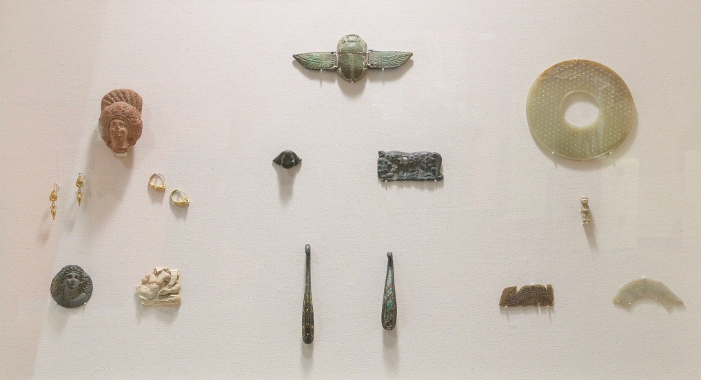 Collection of small objects on white gallery wall: Chinese jade pendants in the shapes of a human figure, a dragon, and a fish can be seen along with Greek and Roman earrings, an Egyptian scarab, and a Sino-Siberian belt plaque