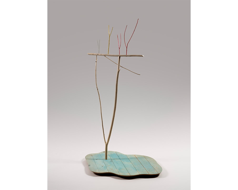 freestanding sculpture; blue painted wood base, two sticks rising up out of the base, smaller colorful sticks balanced on top of those two sticks