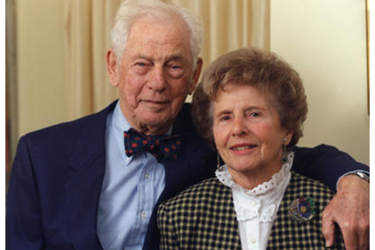 bust portrait of man wearing navy blue suit and bow tie, woman wearing checkered blazer and white shirt; man has his arm around woman
