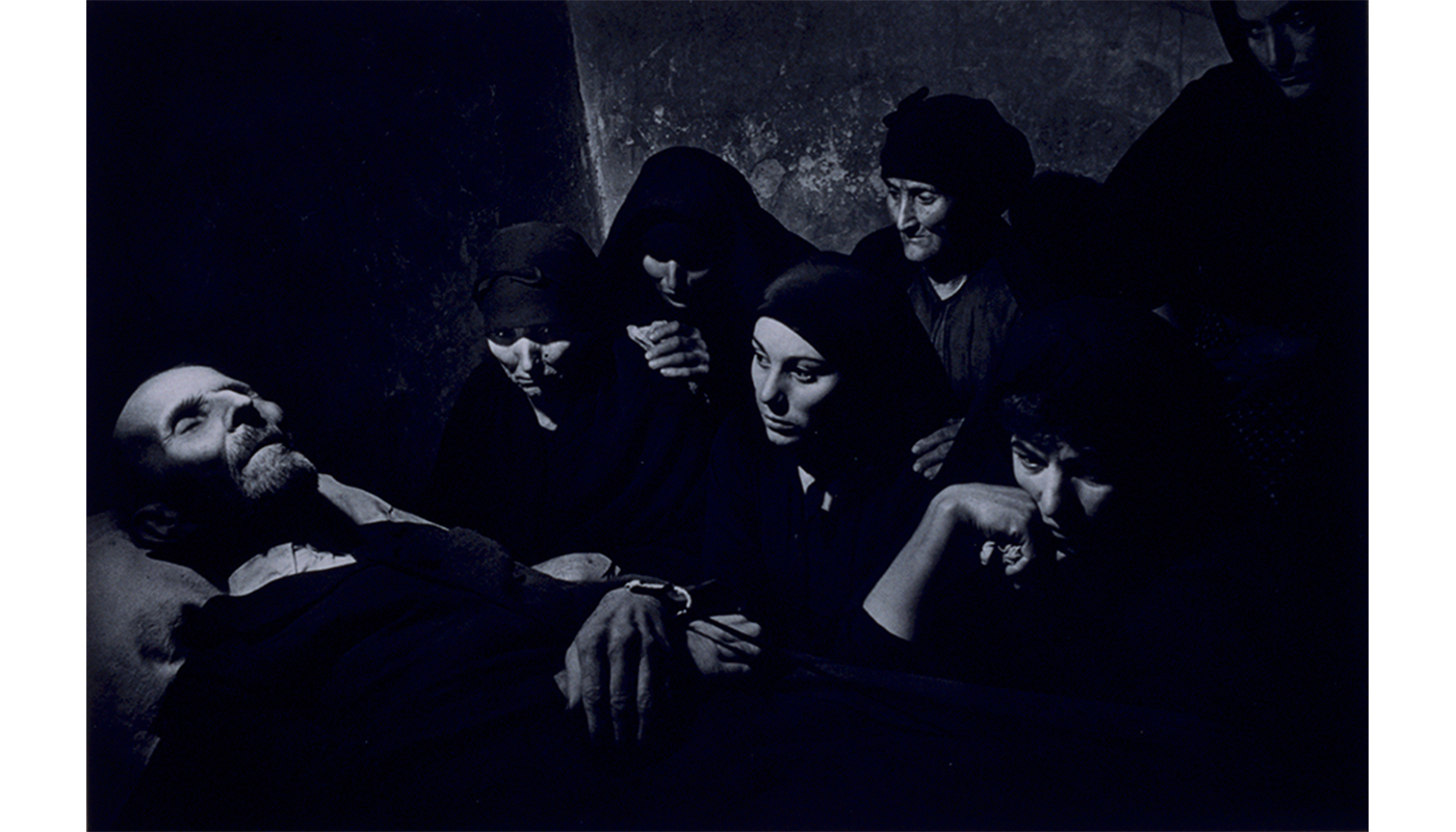 man lying on a bed in the lower left hand corner, a group of women located on the right watch him, they are all in black and the women are wearing black veils