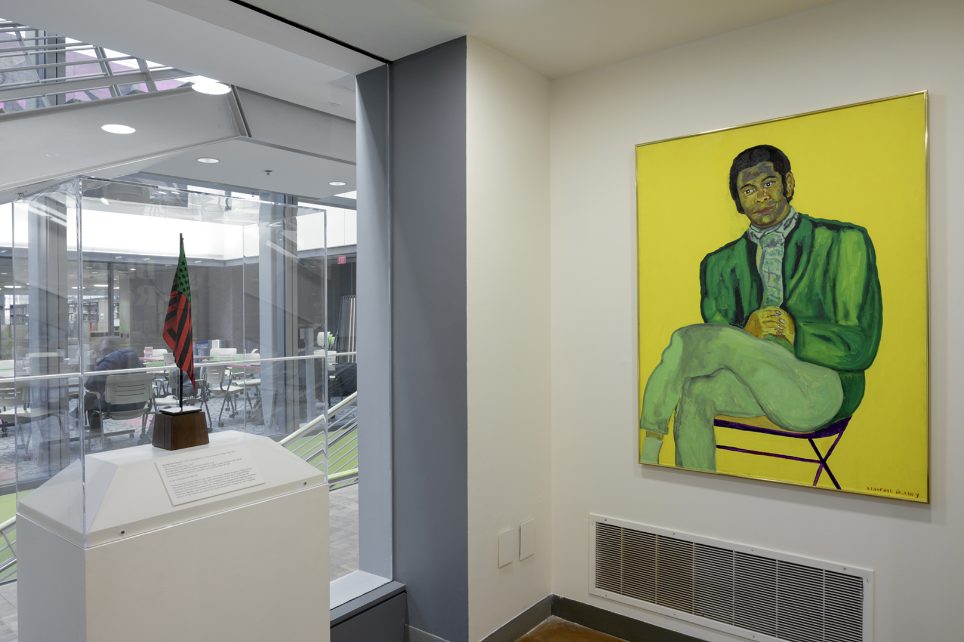 On view in an SCMA gallery next to tall windows to the atrium: left; a small American flag in red, dark blue, and bright green; right: a Black man dressed in all green and seated, legs crossed, on a folding chair on a vibrant yellow background; signed Beauford Delaney in small red block letters at the bottom right.