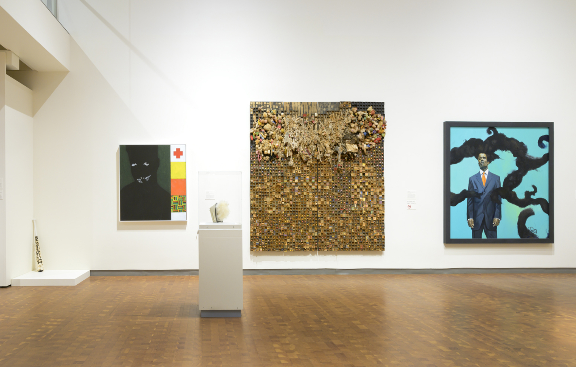 On view in the lower SCMA gallery, from left to right: baseball leaning up against the wall in a corner; painting of a shadowy, toothy-smiling figure next to small squares of color, stripes, and a red cross; an iron emitting soft fabric on a pedestal; a large brown/gold abstract painting made of small squares; a painting of a Black man wearing a blue suit, orange tie standing in front of a blue background with horizontal plumes of black smoke.
