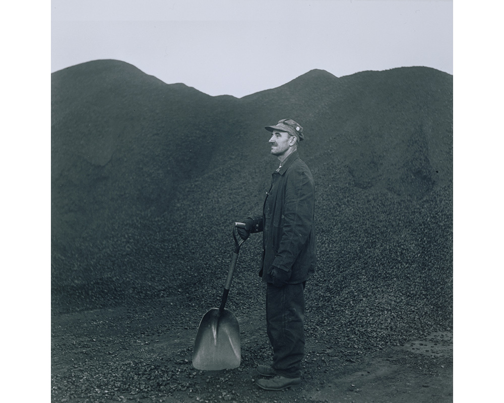 man in work clothes, gloves and hat standing facing proper right in huge pile of coal holding shovel in right hand