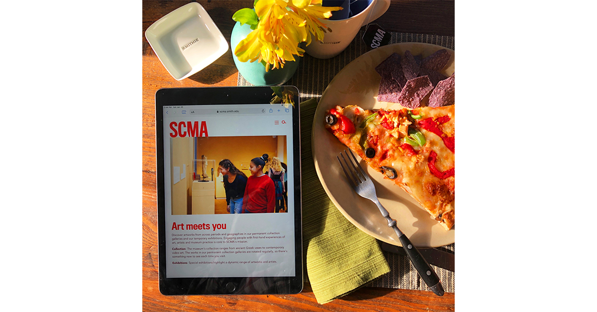 Image of a desk with an IPad opened to SCMA, yellow flowers in a vase, and a slice of pizza on a plate.