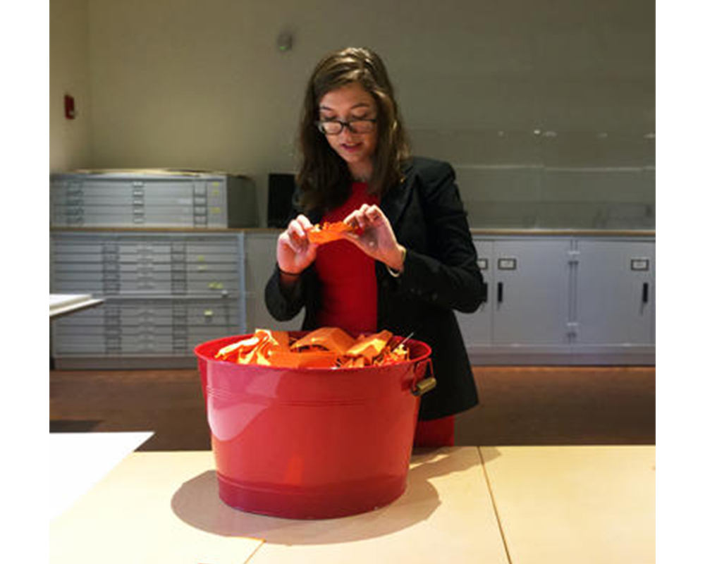 woman with dark hair and glasses picks an orange slip of paper out of a red bucket that contains many orange slips of paper