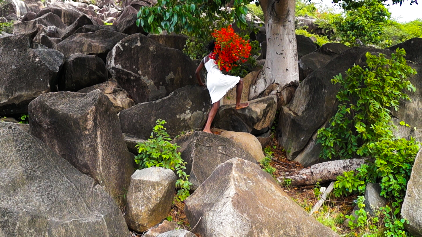 young black girl climbs over large rocks with a large bouquet of red flowers over her shoulder