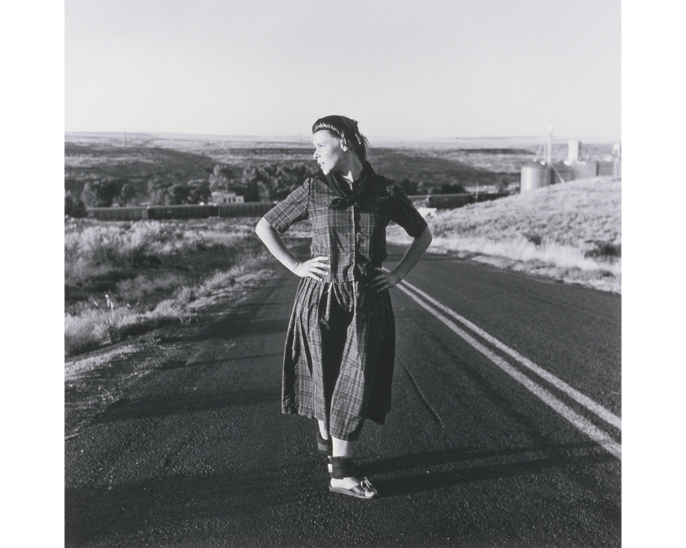 young woman with long plaid dress, sandals, ankle weights, hands on hips, head turned toward proper right standing in land of divided road with fields on either side and parts of buildings visible in low hills at distance