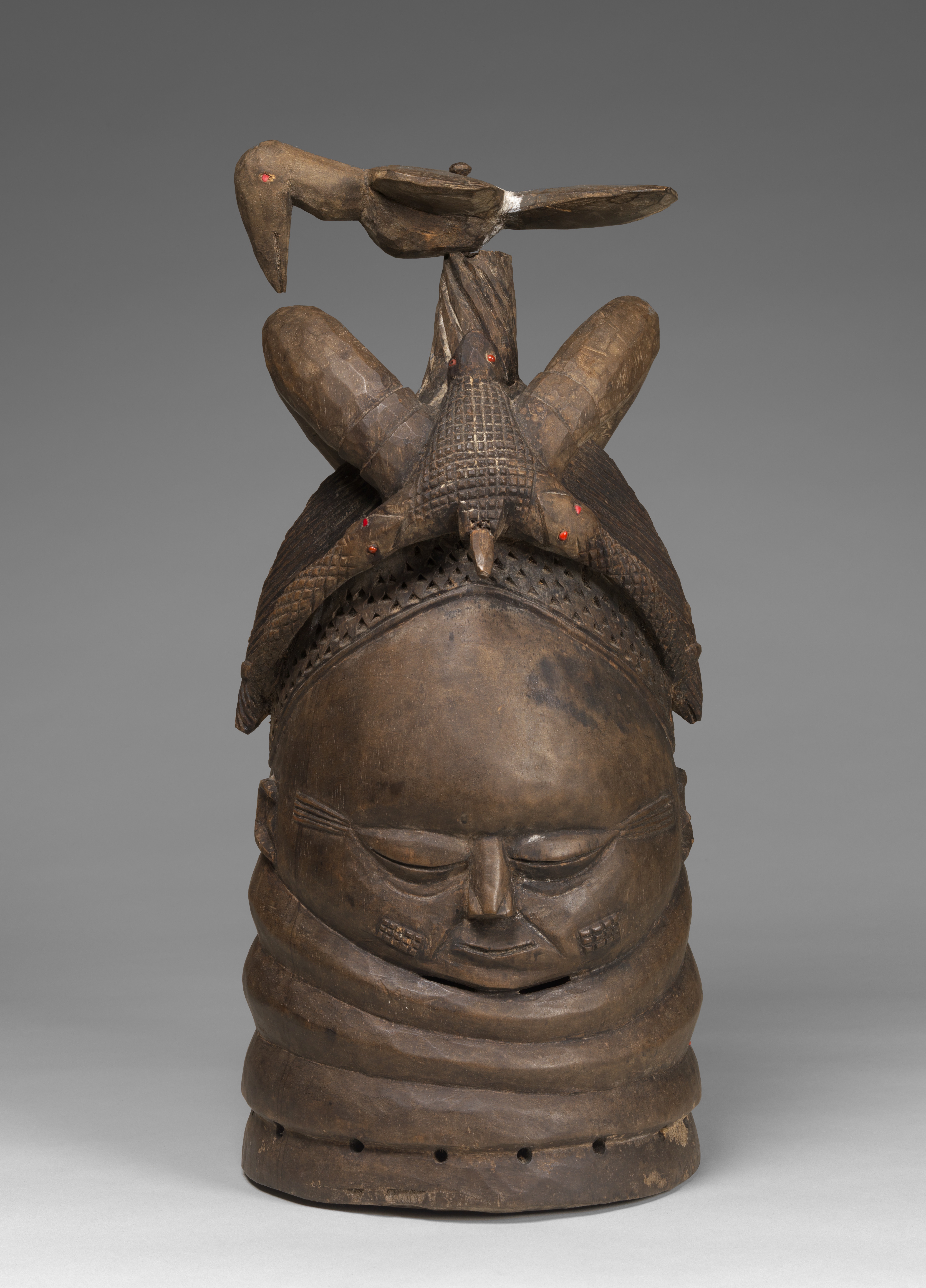 Wood and metal sculpture of a head with closed eyes and a bird shaped headpiece.