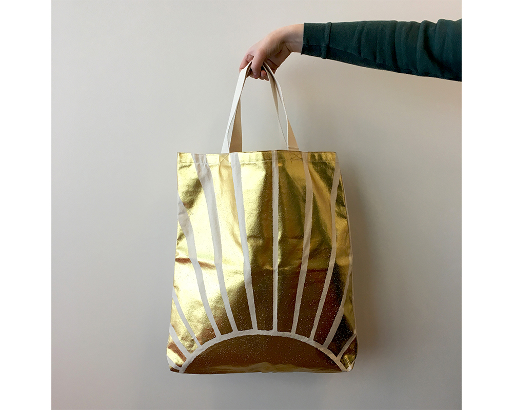 A hand holding a bag, with a golden sun on it, by a strap handle.