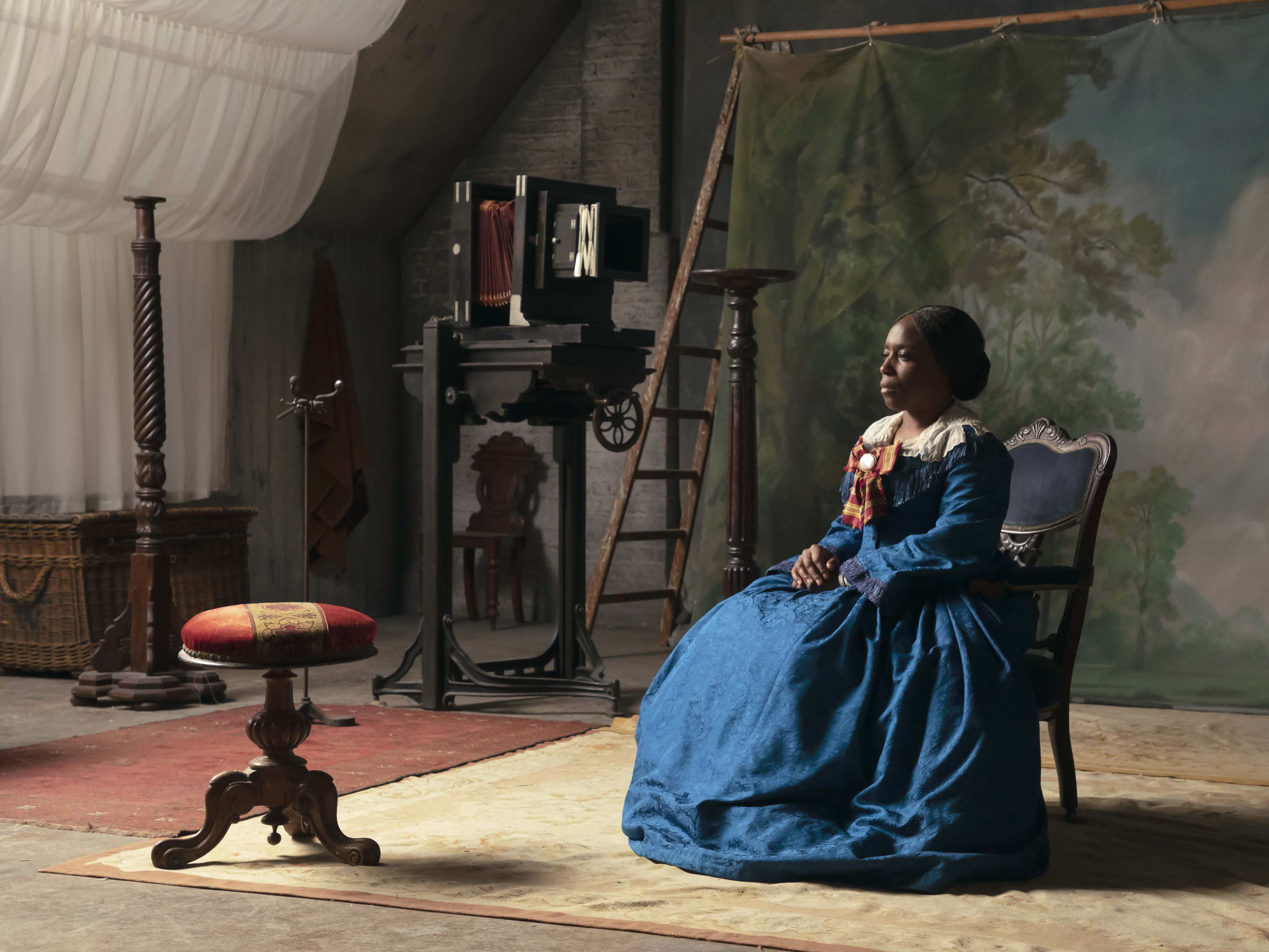 Photograph based on Isaac Julien's Lessons of the Hour of a woman in 19th century floor length saturated blue dress, sitting for a photo portrait
