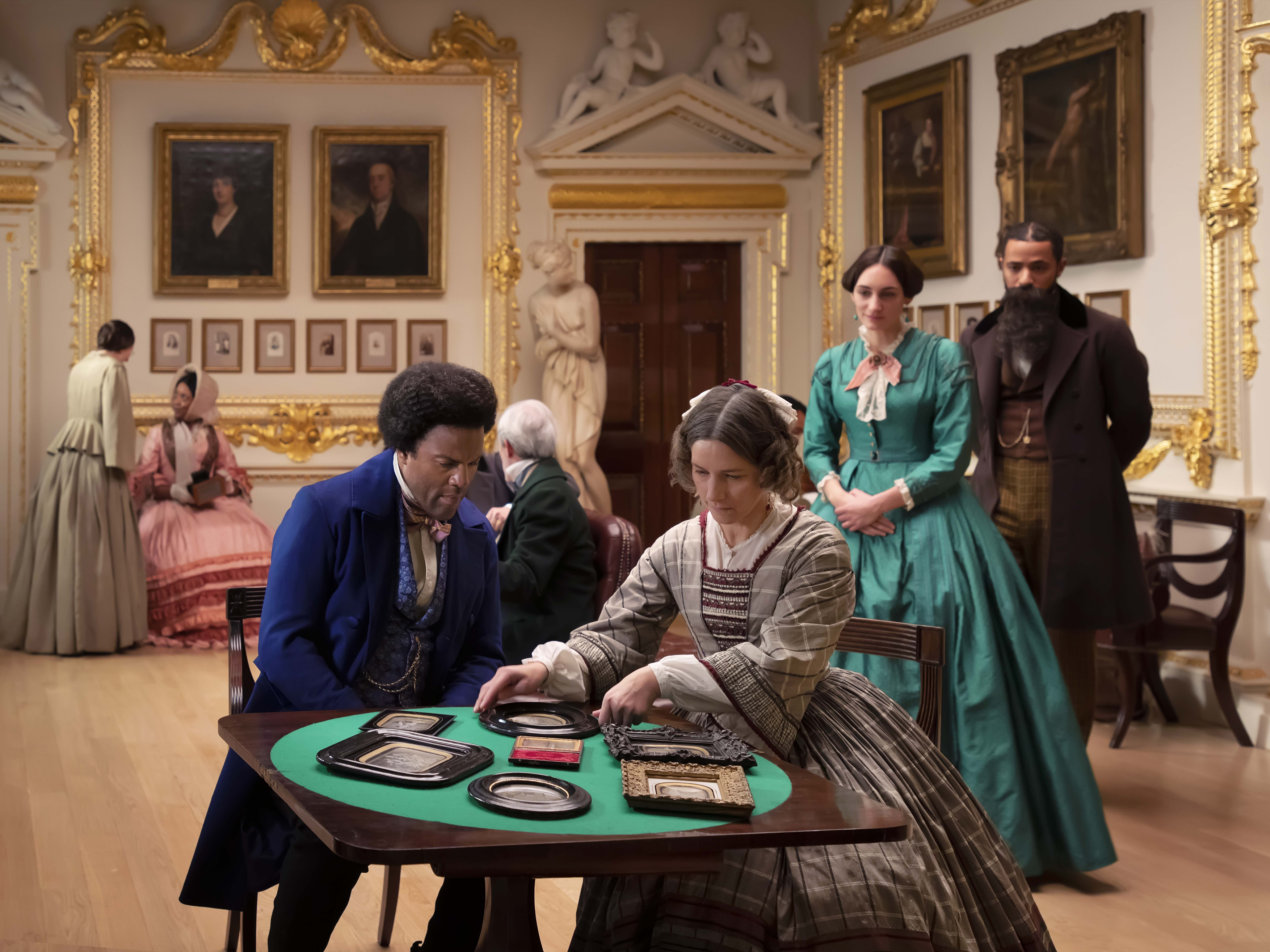 Photograph of Lessons of the Hour of a roomful of people from mid-19th century, a man and woman sitting at a table in foreground looking at photographs