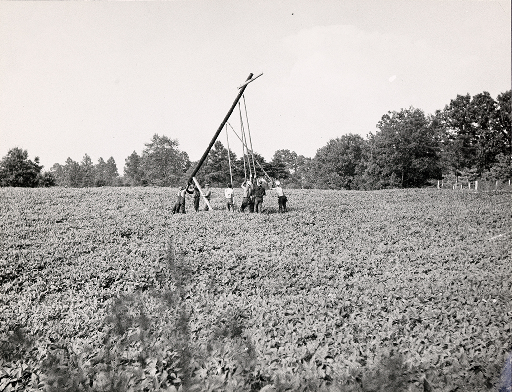 "8 people erecting a wooden pole in a field"