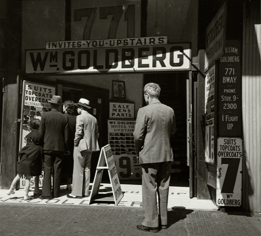 "group of people outside a storefront with a large sign reading "INVITES-YOU-UPSTAIRS / WM. GOLDBERG""